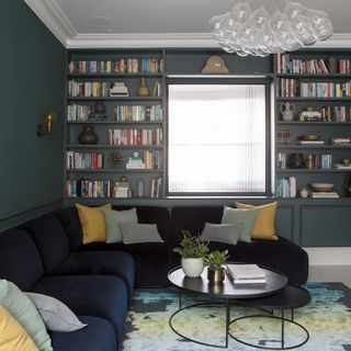 A living room / snug area with built-in bookshelf displaying an array of assorted books, dark blue L-shaped sectional sofa and blue/yellow area rug with abstract motif and statement chandelier light fixture