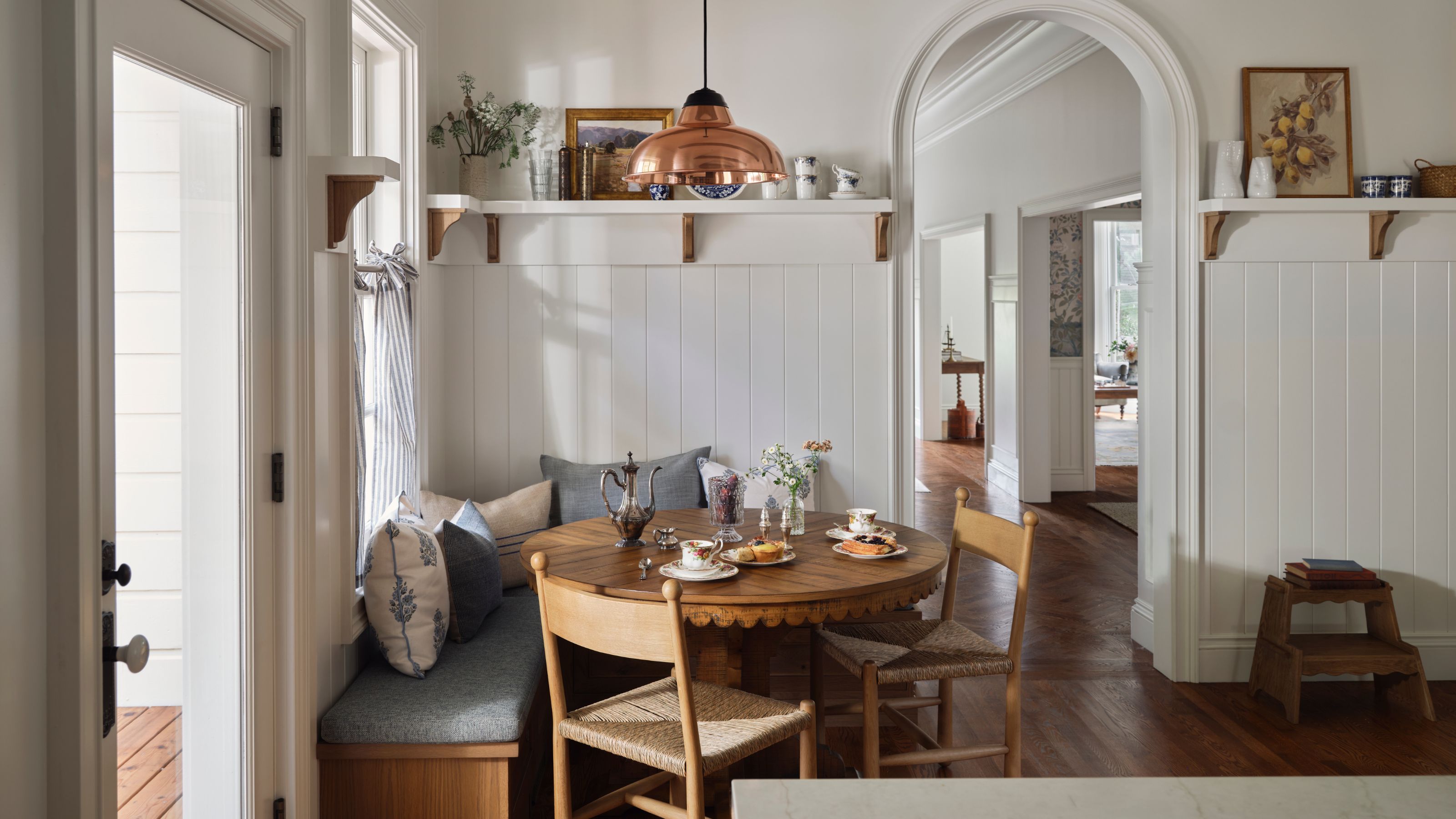 a large white painted paneled kitchen with a breakfast nook styled with banquette seat, round table and oak chairs with shelves displaying vintage finds above