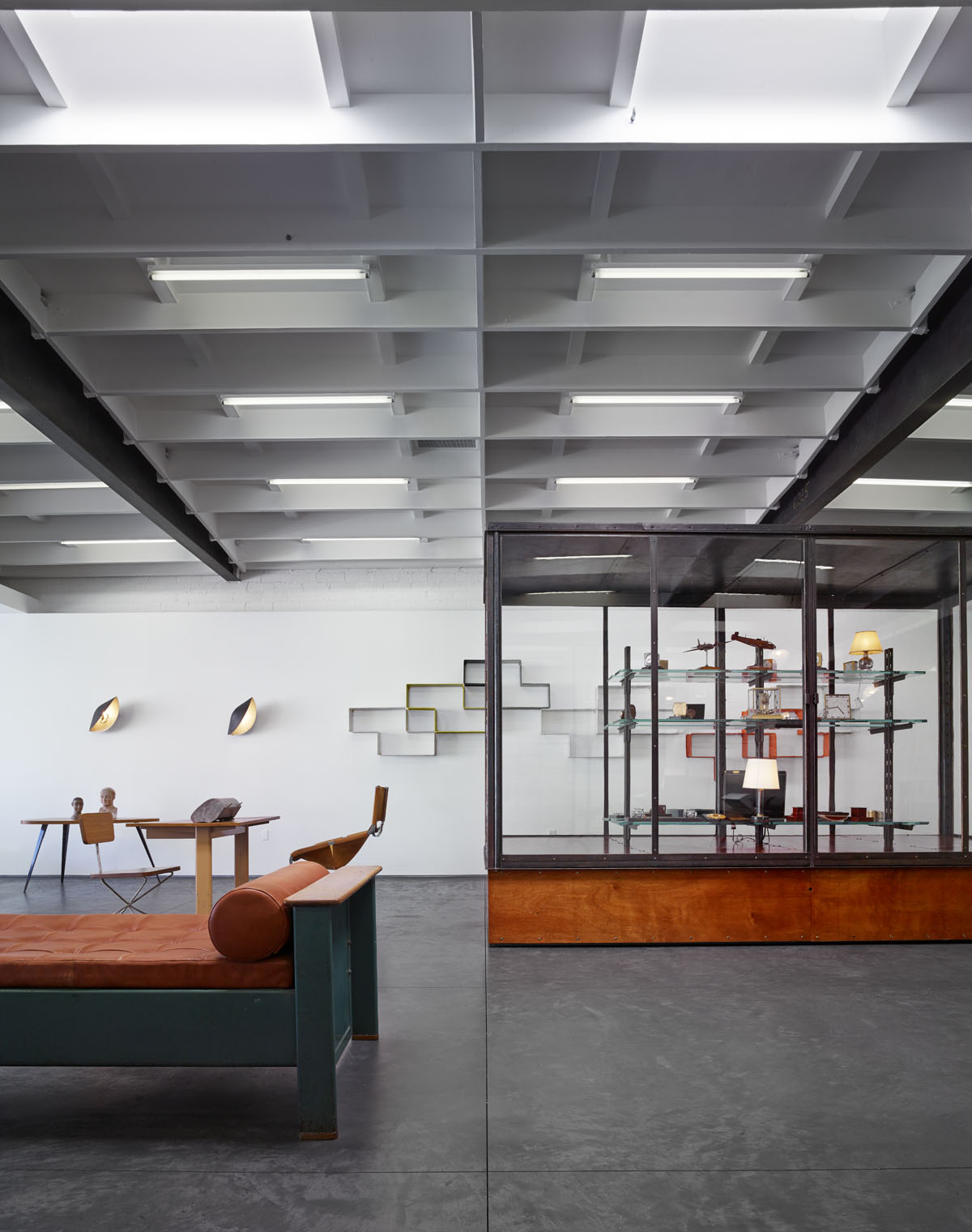 A dramatically designed gallery space with retro coffered ceilings is filled with industrial-style furniture, including a velvety, geometric single bed, wooden table sets, modular metallic shelves, and a large library.