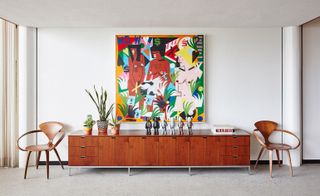 Large painting on a wall behind a large sideboard flanked by 2 wooden arm chairs