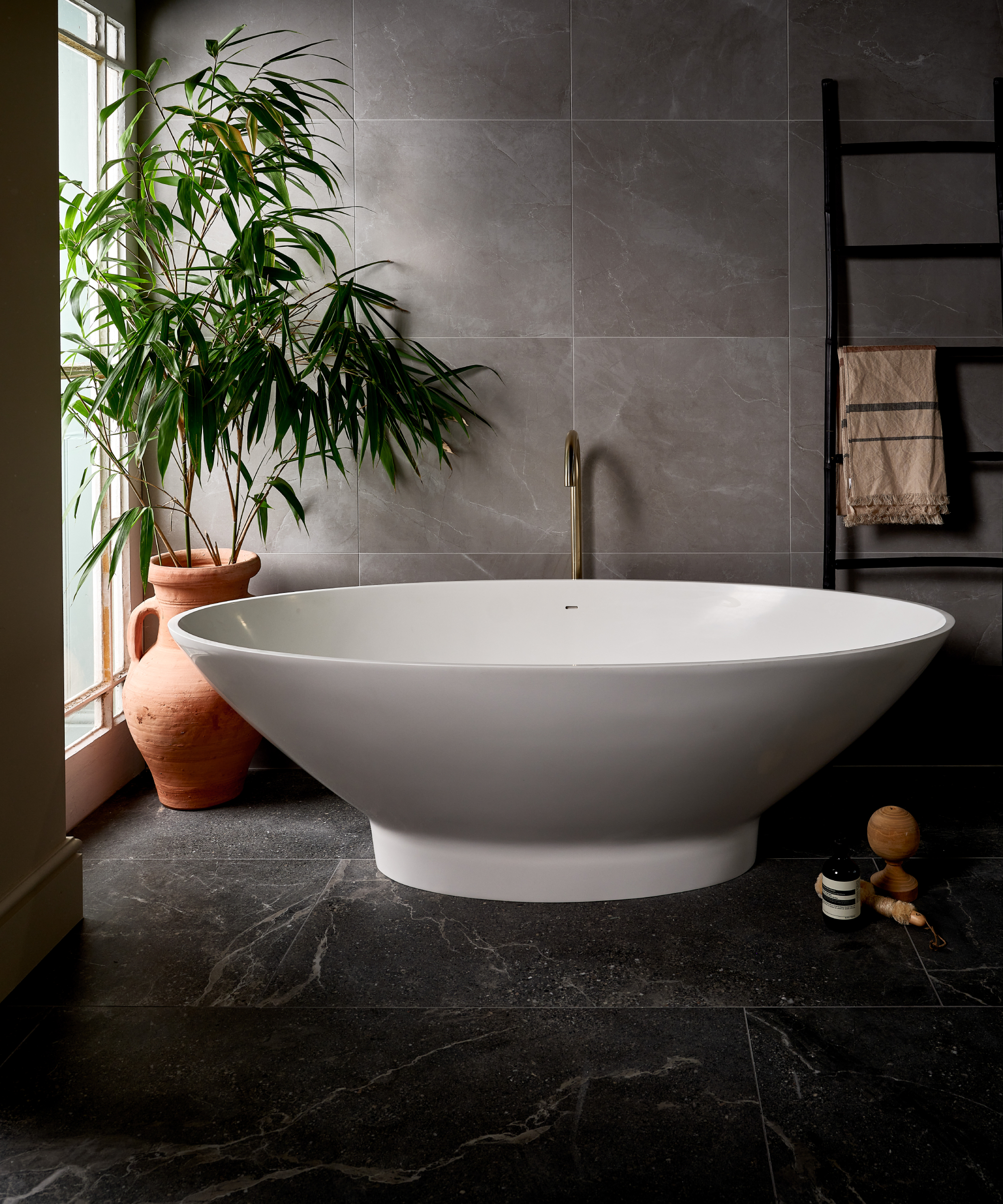 white modern freestanding bath in bathroom with dark veined marble floor tiles and grey wall tiles, black towel radiator, green plant in corner next to large window