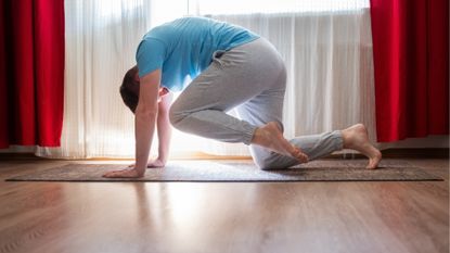 A man performs the bird-dog exercise on a yoga mat in a room with wooden flooring. He is on his hands and knees, with his left knee lifted up towards his head. He pushes against the floor to round his back.