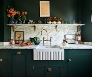 A dark green kitchen with marble countertops and backsplash, gold hardware, and a fluted detail sink