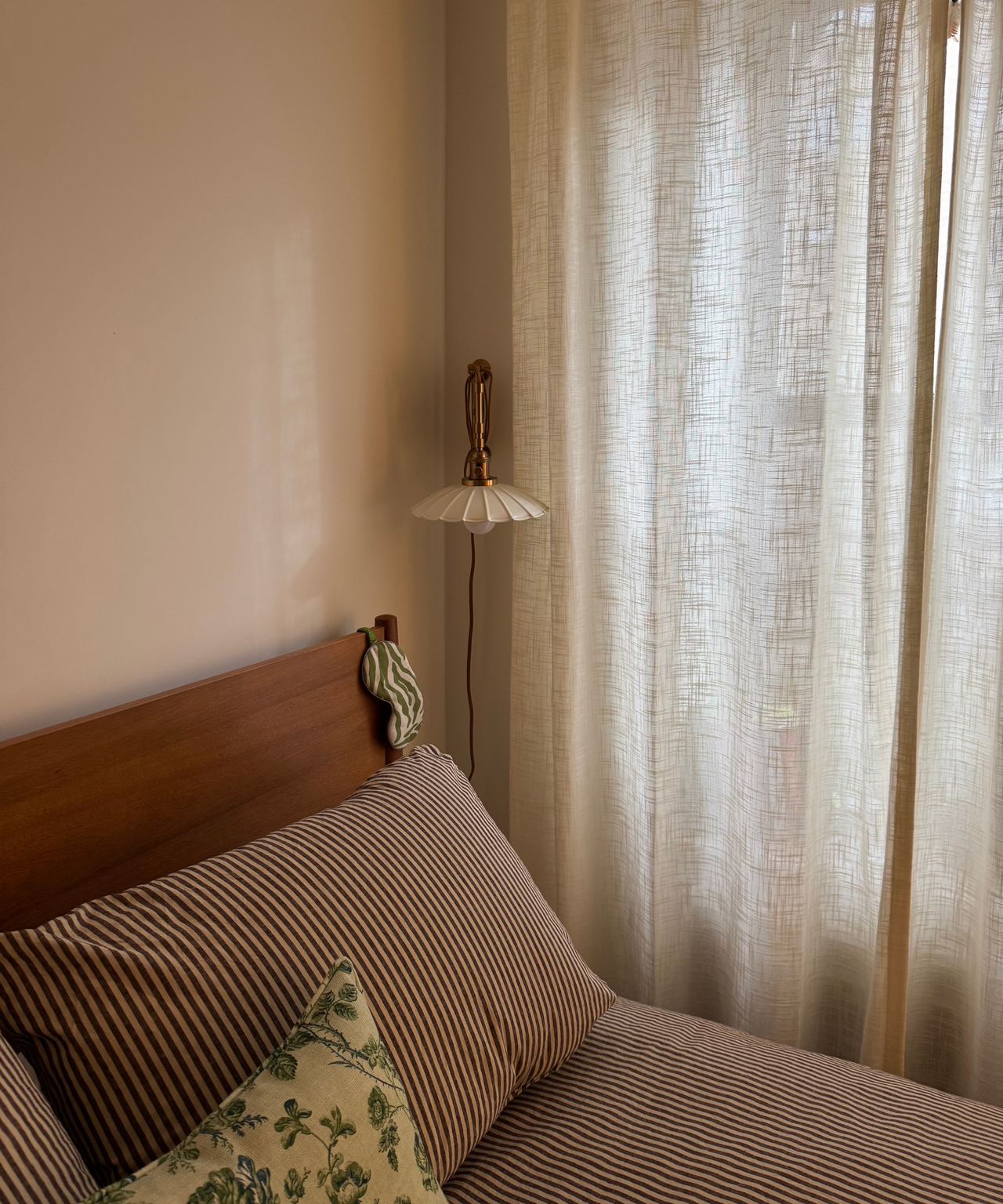 a bedroom with ivory curtains, a wooden bed frame, striped bedding, and a plug in wall sconce