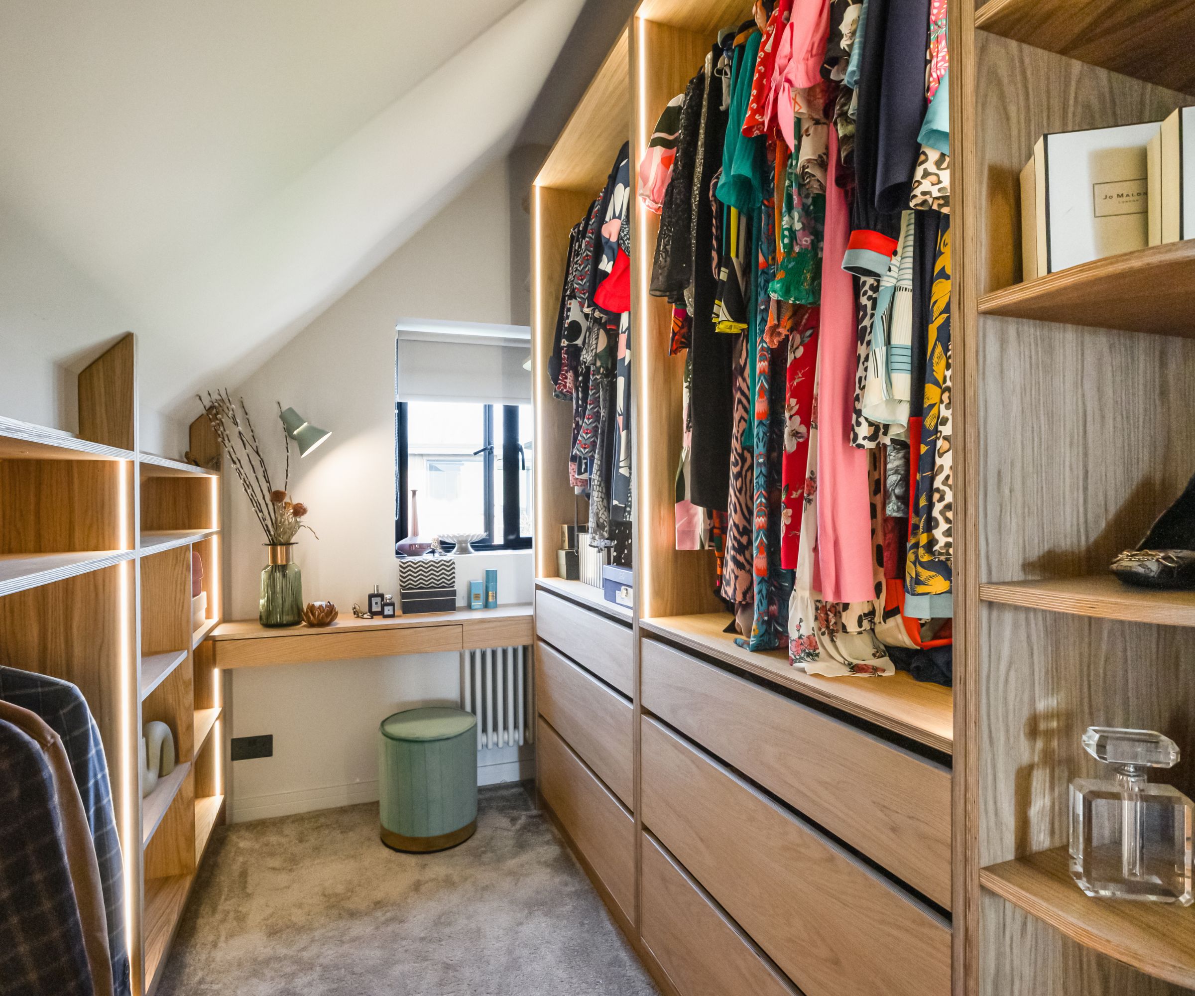 A narrow walk-in wardrobe beneath a sloping loft conversion ceiling. One side holds colourful dresses on a hanging rail above wide wooden drawers; the other features illuminated open shelving. At the end, a small desk and round green stool sit below a window.