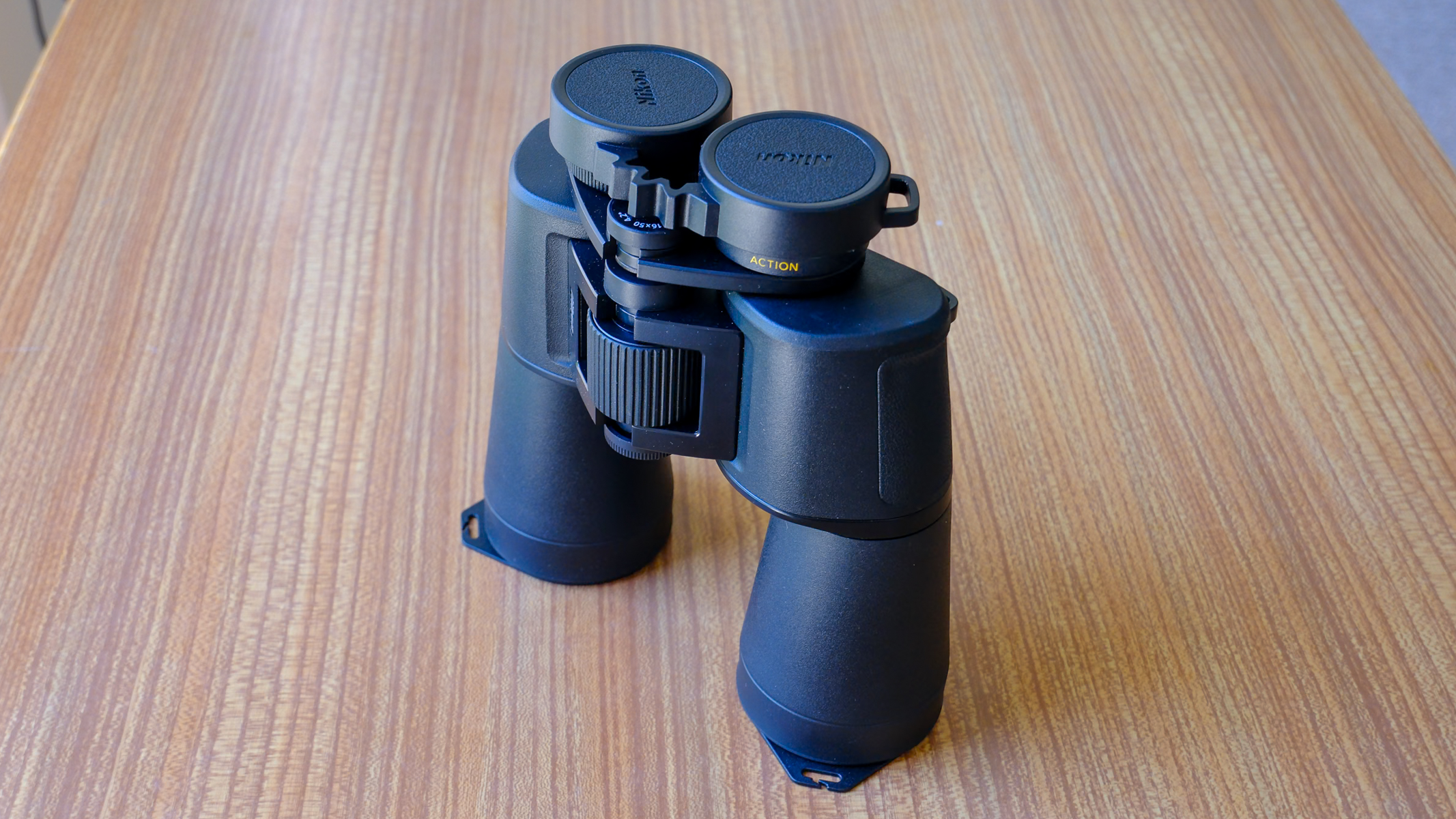 The Nikon Action 16x50 binoculars stood up on a wooden table with eyecup covers and focus dial visible.