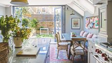 Bright kitchen and dining area with large plants in vases, kitchen island, dining table with cushioned chairs, skylights, and open doors leading to small, sunny, outdoor area.