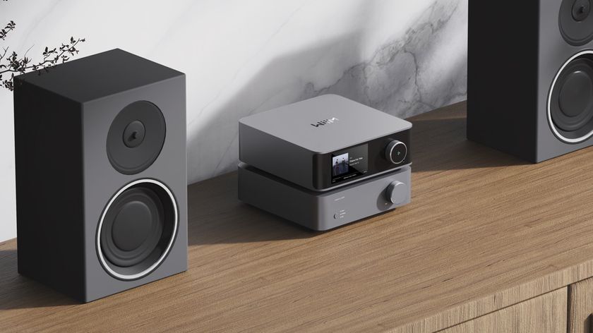 A grey WiiM Vibelink Amp on a wooden cabinet between two bookshelf speakers.