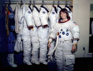 a woman in a white spacesuit stands in front of a rack with six white spacesuits hanging from it.