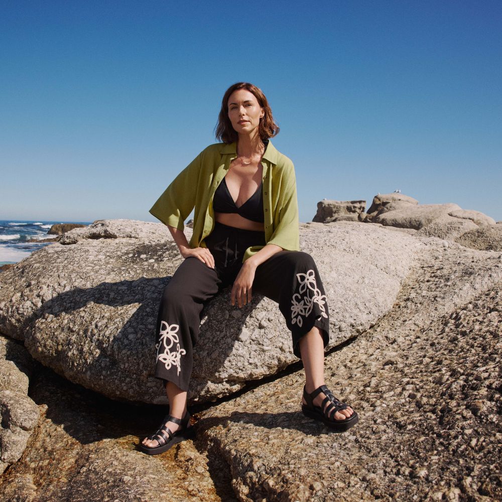Model sat on a boulder on the beach wearing black trousers with floral embellishment, a black tirangle bikini top and green short sleeve shirt sold at JD Williams