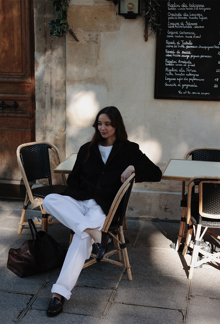 A woman sitting at a cafe wearing a black blazer, white t-shirt, white baggy jeans, black loafers, and a black bowler bag
