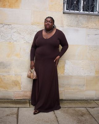 influencer and creative Abisola Omole poses on a sidewalk in Copenhagen wearing a long-sleeve brown maxi dress, small snakeskin bag, and brown suede Manolo Blahnik mule heels