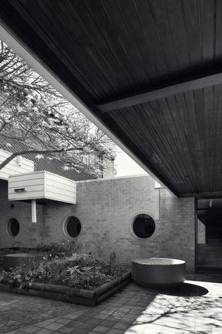 A black and white photo of a courtyard with a plant area, brick walls with round windows and brick flooring.