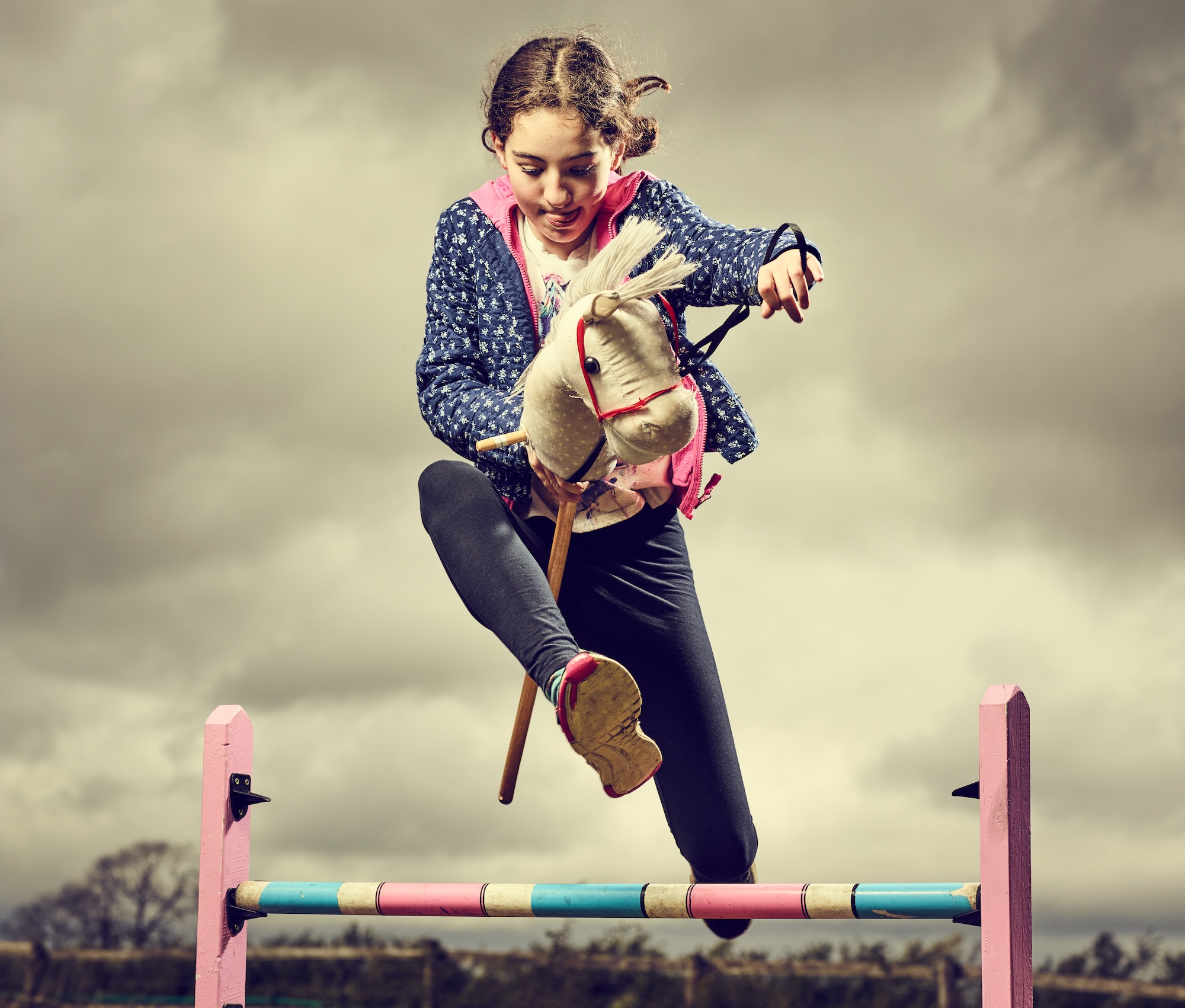 Young girl hobby horsing over a jump, with a cloudy sky above