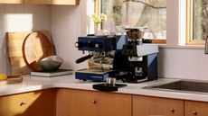Image of a navy blue La Marzocco coffee machine next to a matching coffee grinder on a white countertop with wooden cabinetry in a white tile kitchen.