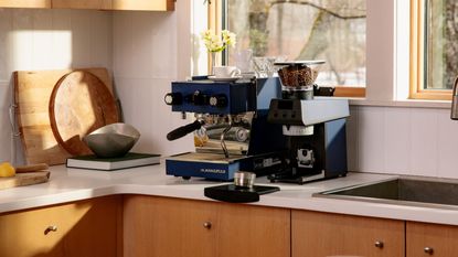 Image of a navy blue La Marzocco coffee machine next to a matching coffee grinder on a white countertop with wooden cabinetry in a white tile kitchen.
