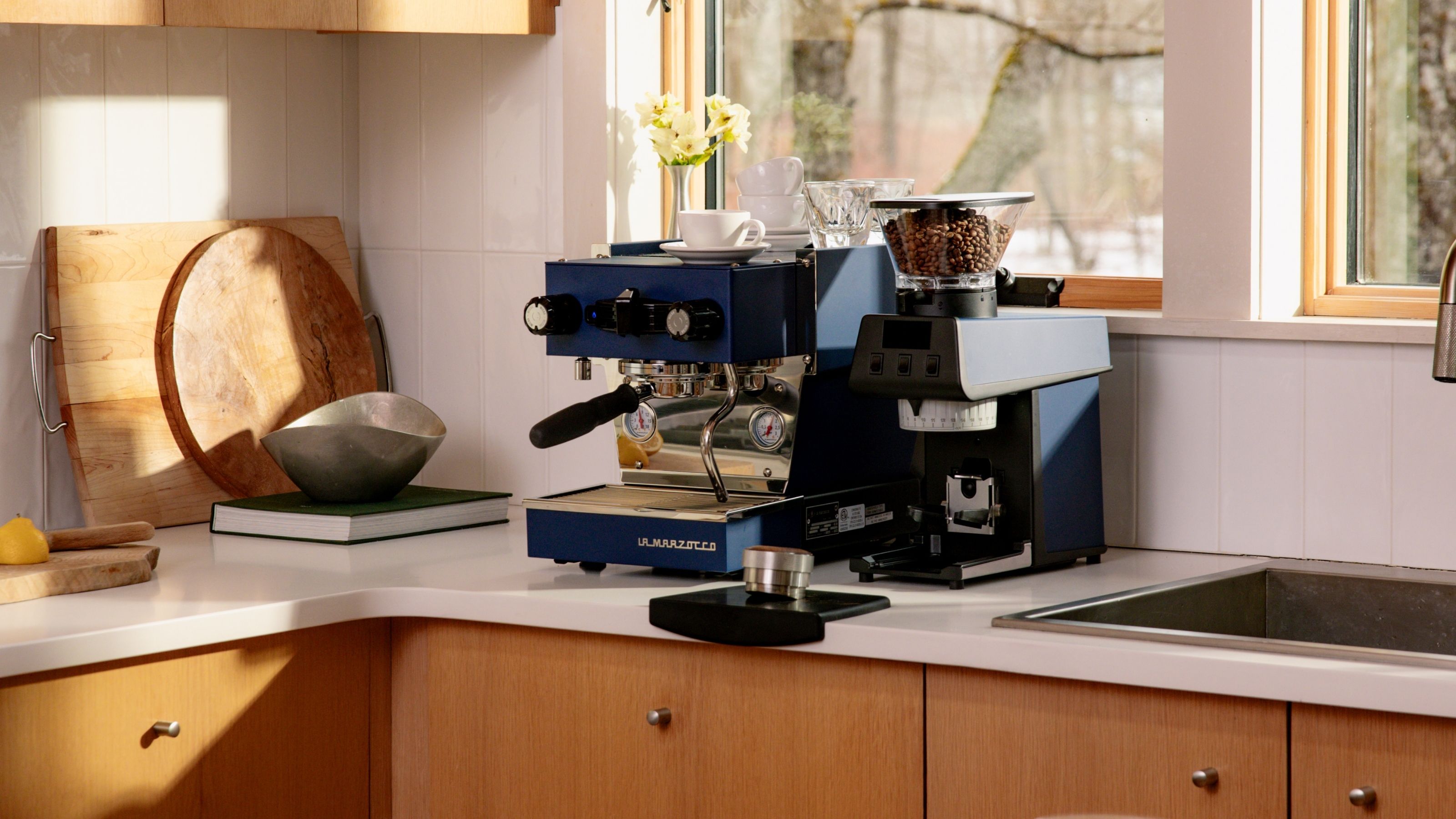 Image of a navy blue La Marzocco coffee machine next to a matching coffee grinder on a white countertop with wooden cabinetry in a white tile kitchen.