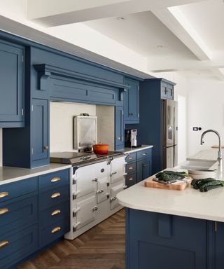 A kitchen with dark blue cabinets and drawers, a white oven with a bright orange pot, and a kitchen island to the right with a silver sink, chopping board, and a white worktop
