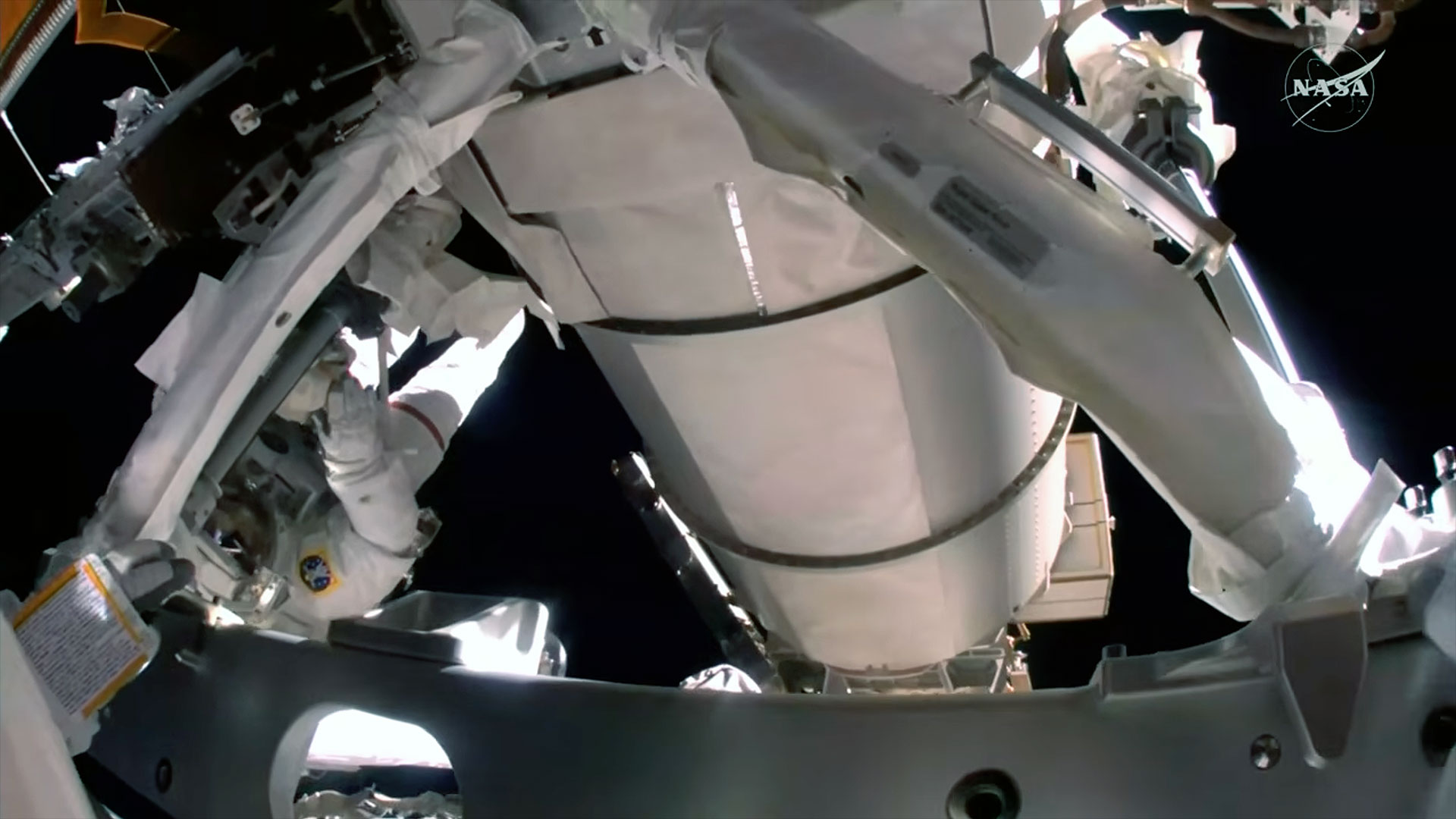 A view from an astronaut's helmet-mounted camera shows the worksite for a spacewalk and a fellow spacesuited astronaut nearby.