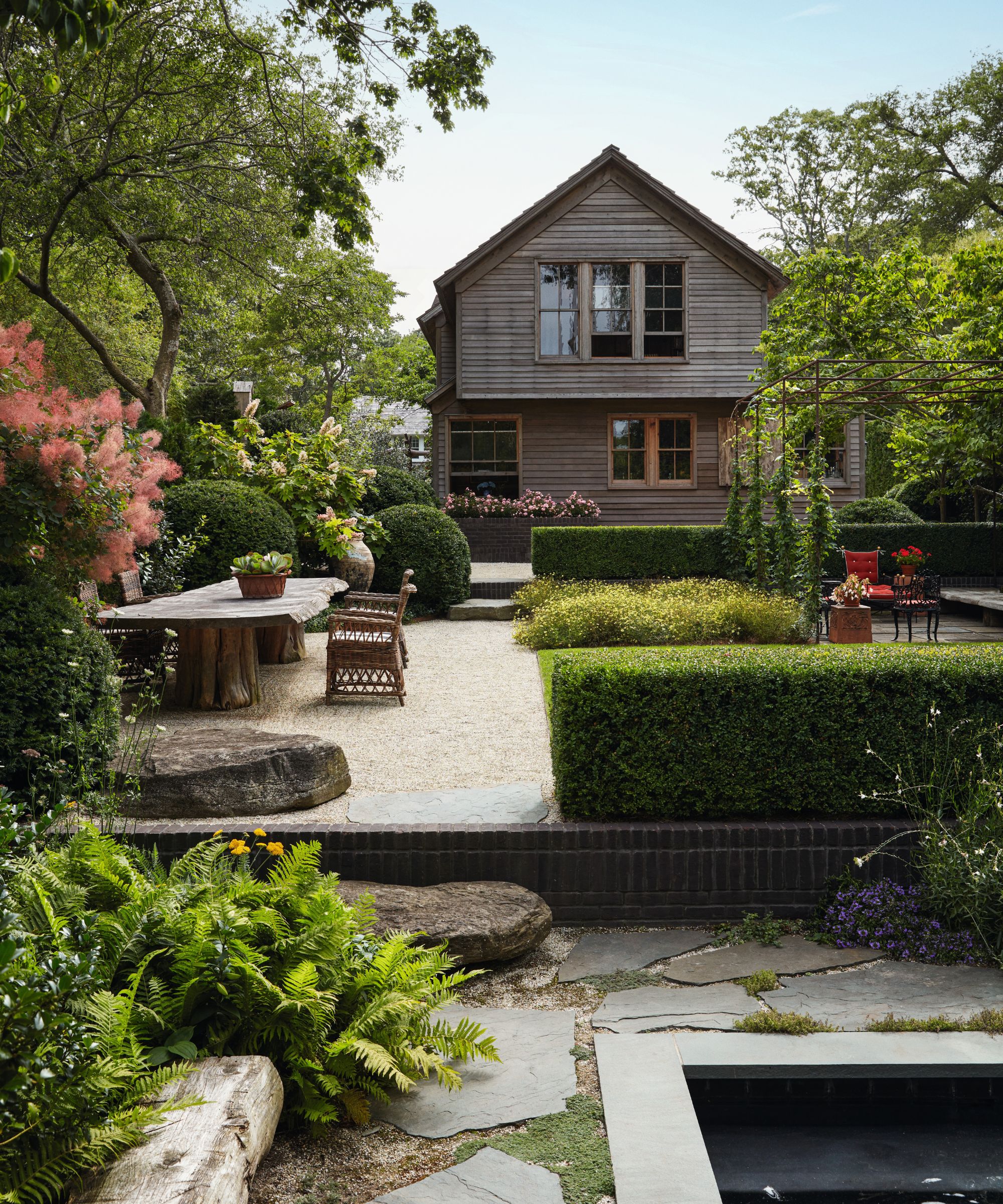 A lush, tiered backyard garden with manicured hedges, stone pathways, and a large outdoor wooden dining table. In the background stands a weathered two-story grey shingle house surrounded by mature trees.