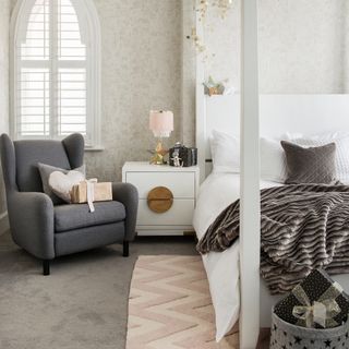 White bed, white bedside table and dark grey armchair on top of dark grey carpet and neutral walls