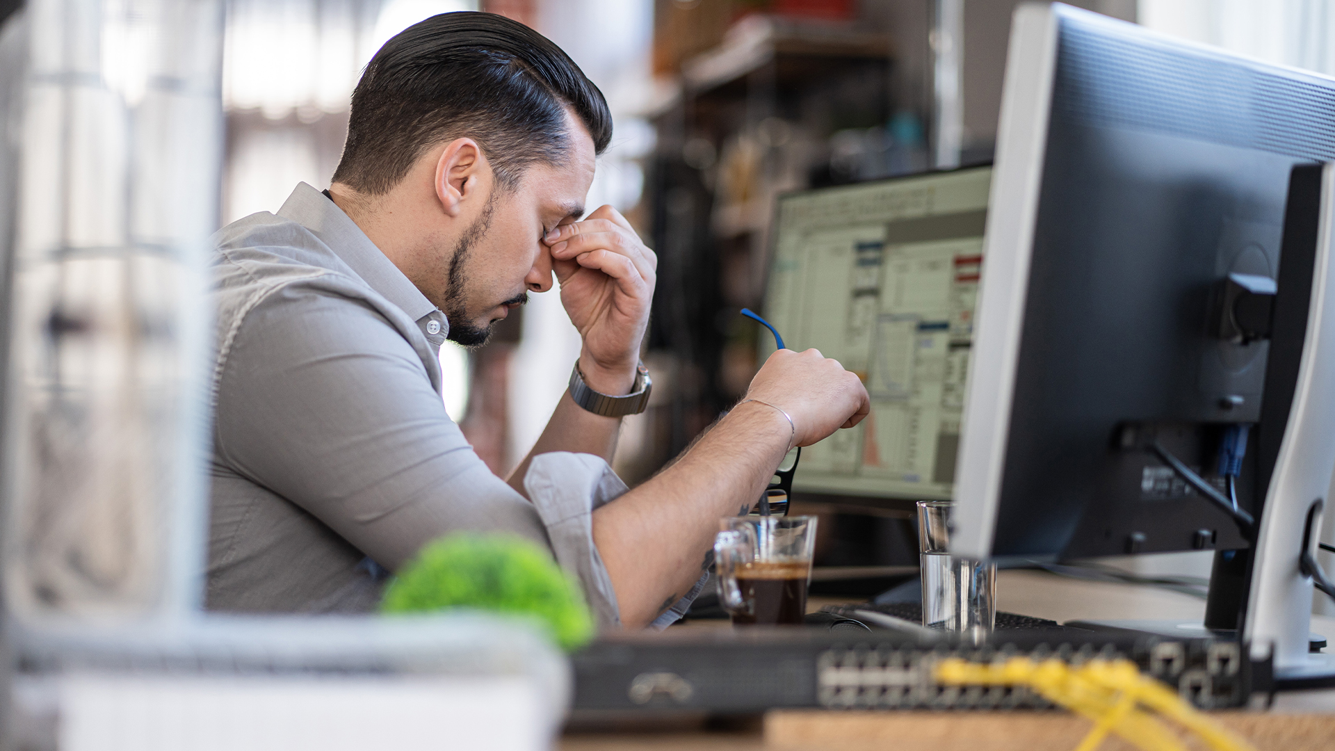 Male cybersecurity worker at desk in office space holding head in signs, signifying stress and burnout. 