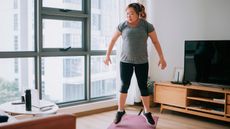 Woman exercising in living room, in midair and looking at laptop on coffee table