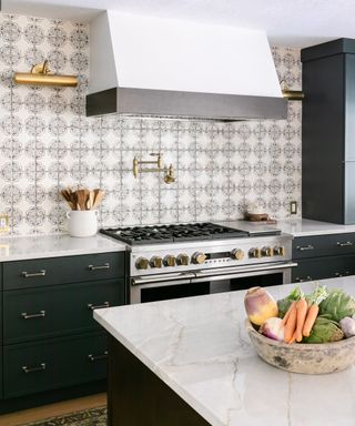 A kitchen with a marble kitchen island with an antique bowl filled with carrots, artichokes, and swedes, with a kitchen with black and white circle splashback behind it with a hood, silver oven, and black cabinets