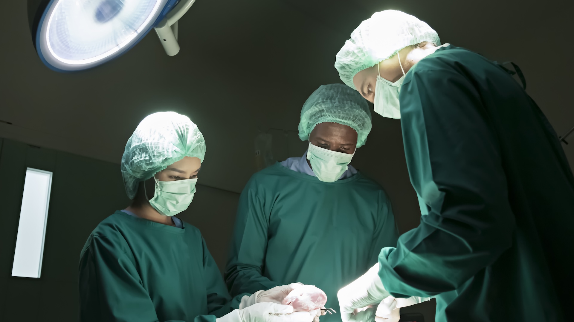 Low angle photo of three doctors in scrubs, gloves, and masks operating on a patient who is out of the frame. 