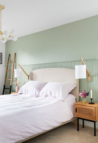 A pale sage green bedroom with paneling and white bedside lampshades and ladder