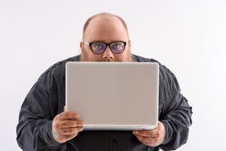 A man wearing glasses holds a laptop up to gingerly cover the lower half of his face.