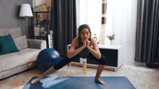 woman doing a side lunge in a living room setting in front of the camera on a blue exercise mat. there's a sofa, window and large exercise ball behind her. 