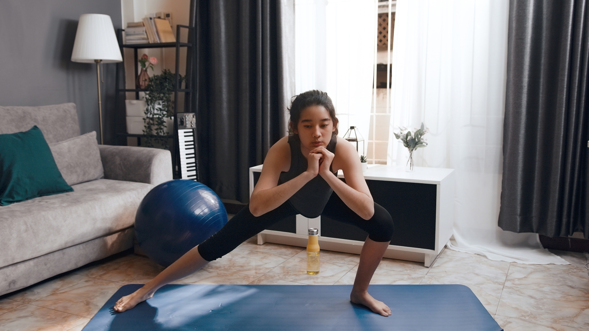 woman doing a side lunge in a living room setting in front of the camera on a blue exercise mat. there&#039;s a sofa, window and large exercise ball behind her. 