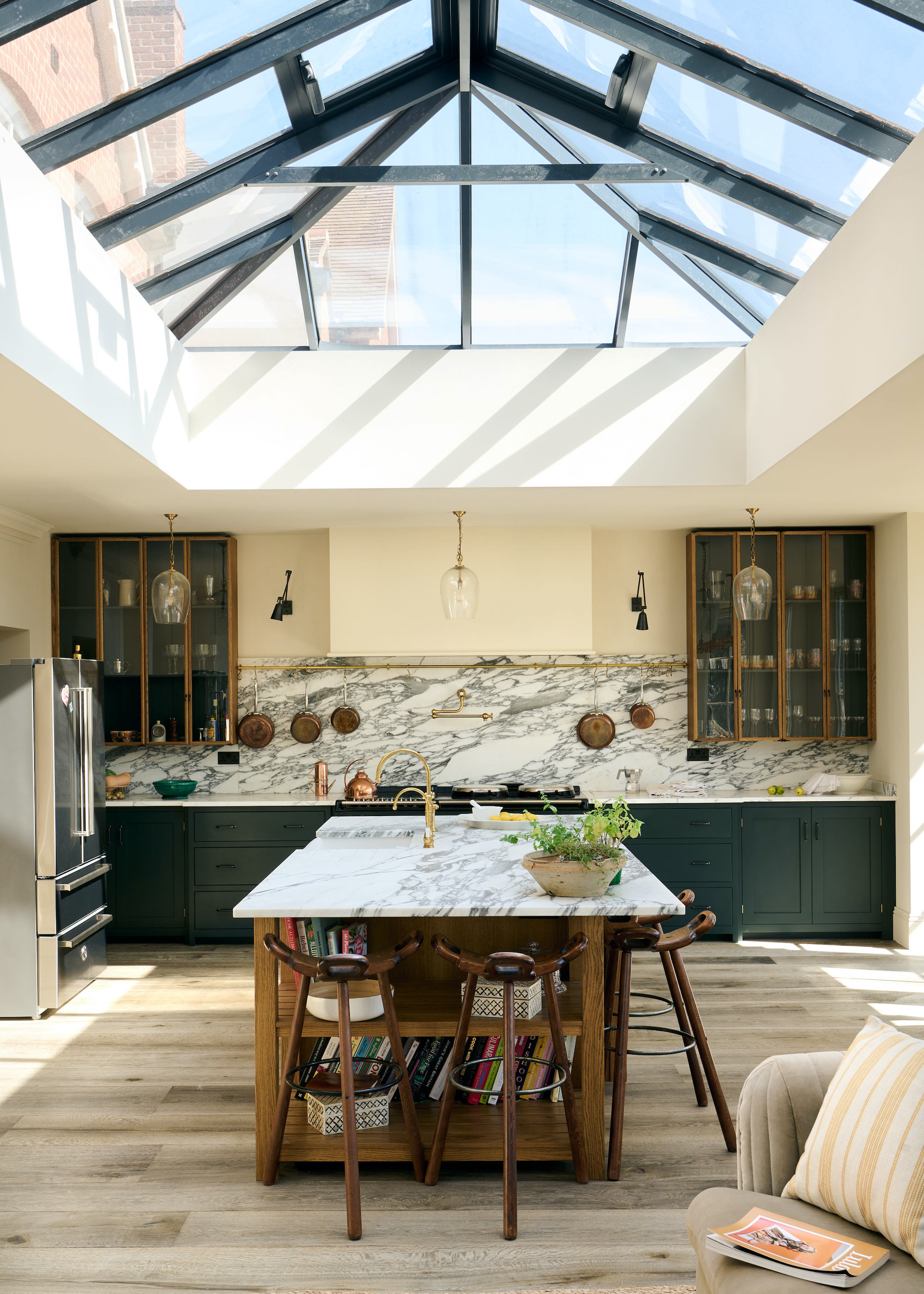 A large kitchen island sits in the middle of a kitchen extension. The lower cabinets of this kitchen are painted green. The upper cabinets on each side are glass-fronted.