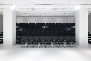 Pictured: rows of black chairs inside an auditorium-like space.