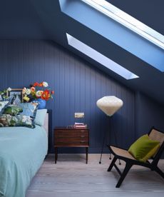 Blue bedroom with slanted roof and ceiling windows, mid century wood and rattan chair with green pillow, walnut nightstand drawers and paneled blue walls