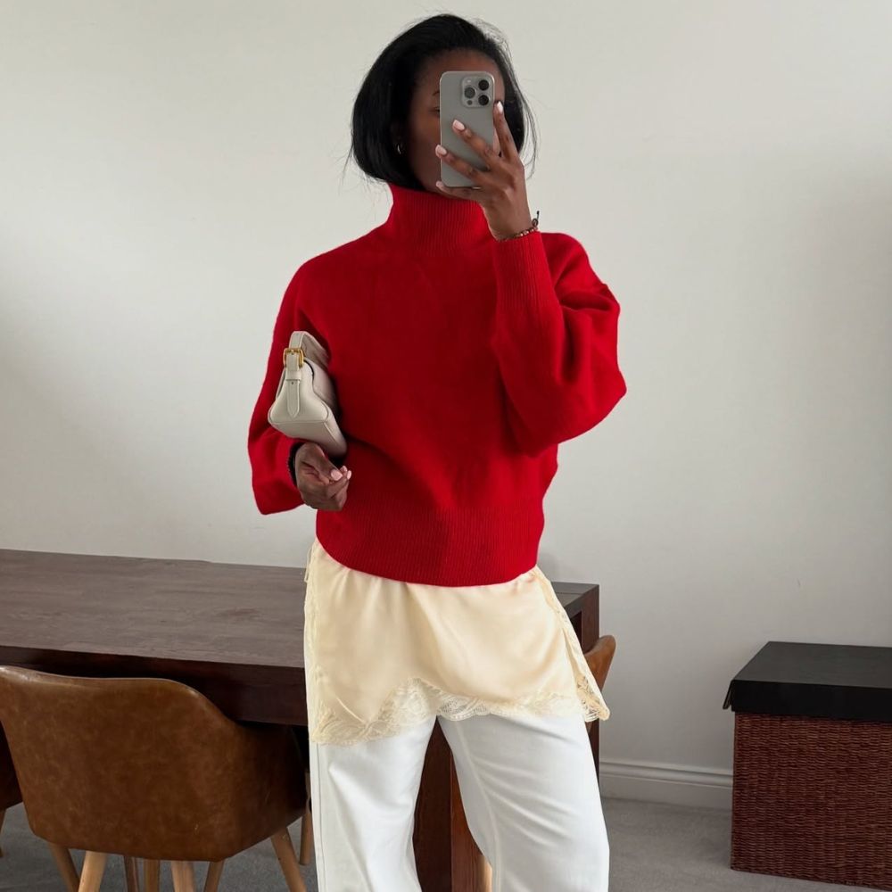 Influencer wears a long top with a lace trim over white jeans and underneath a red knit.