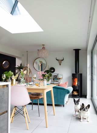 Tile slate light gray flooring in modern living room with Scandi-style pine table, pink dining chairs and other accents plus teal velour sofa, chiminea and pugs.