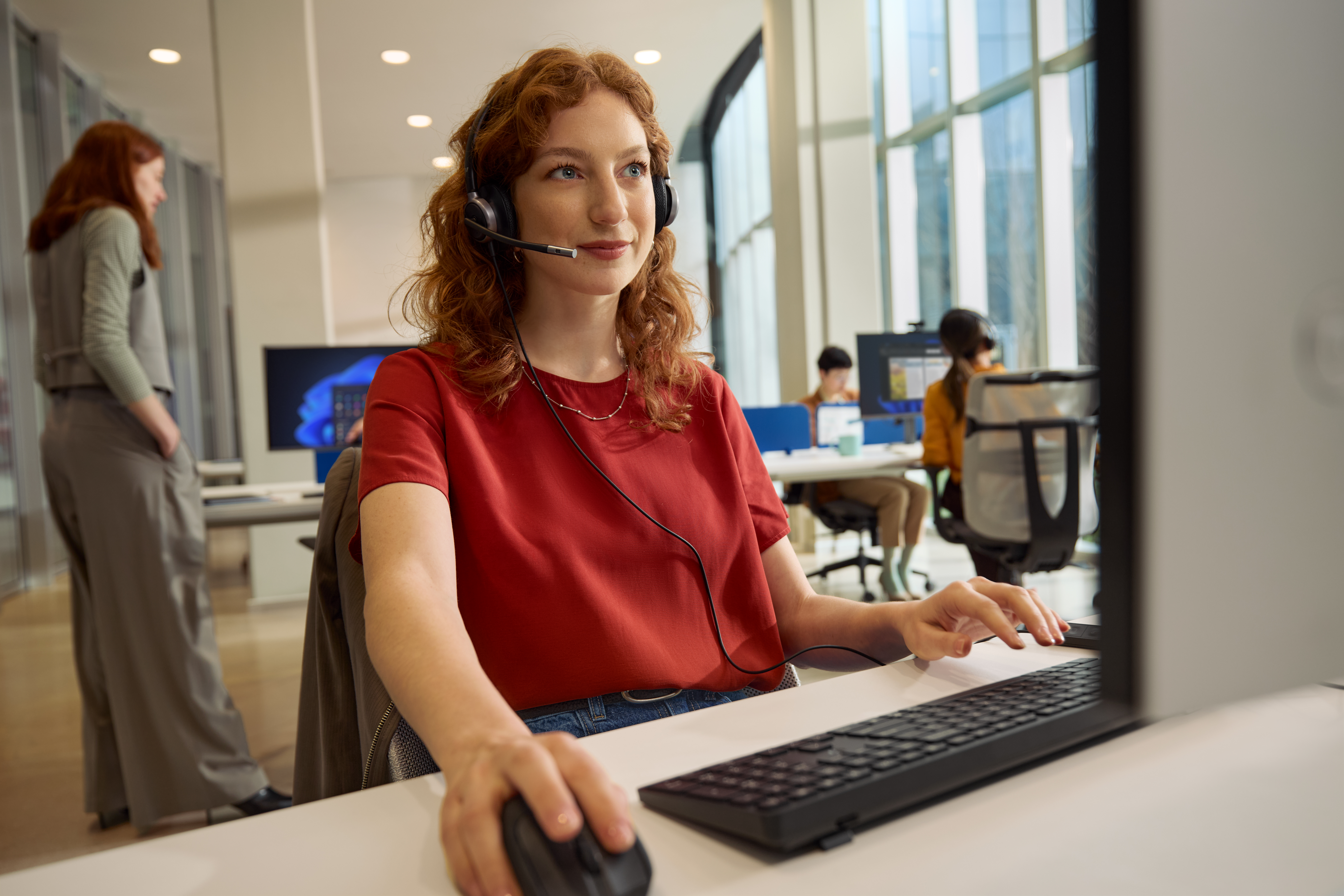 A woman with red hair using a headset while on a desktop computer scrolling her mouse.