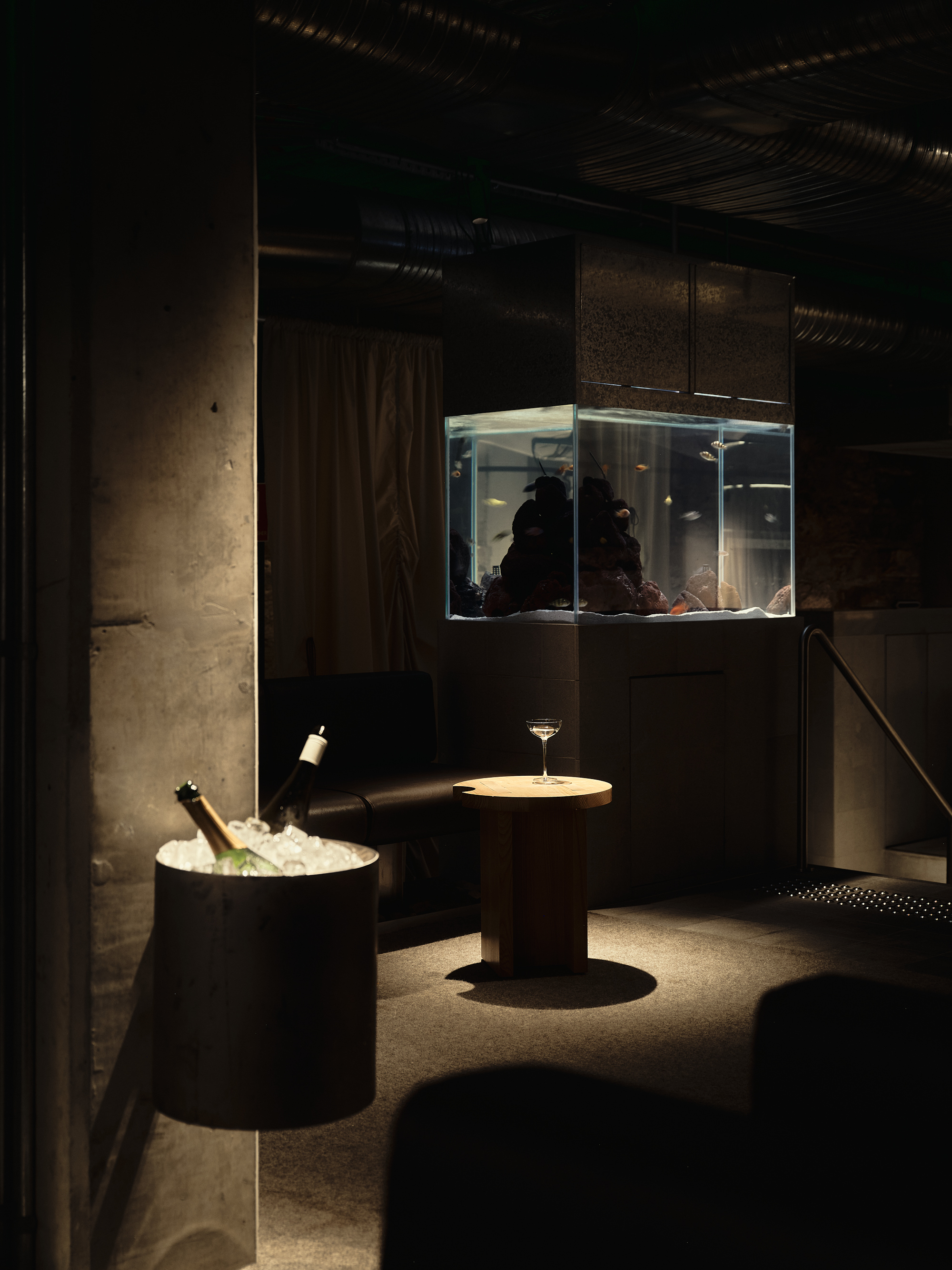 a dark room with concrete walls, a built-in fish tank, a wine glass on a small table, and a bucket of ice holding two wine glasses
