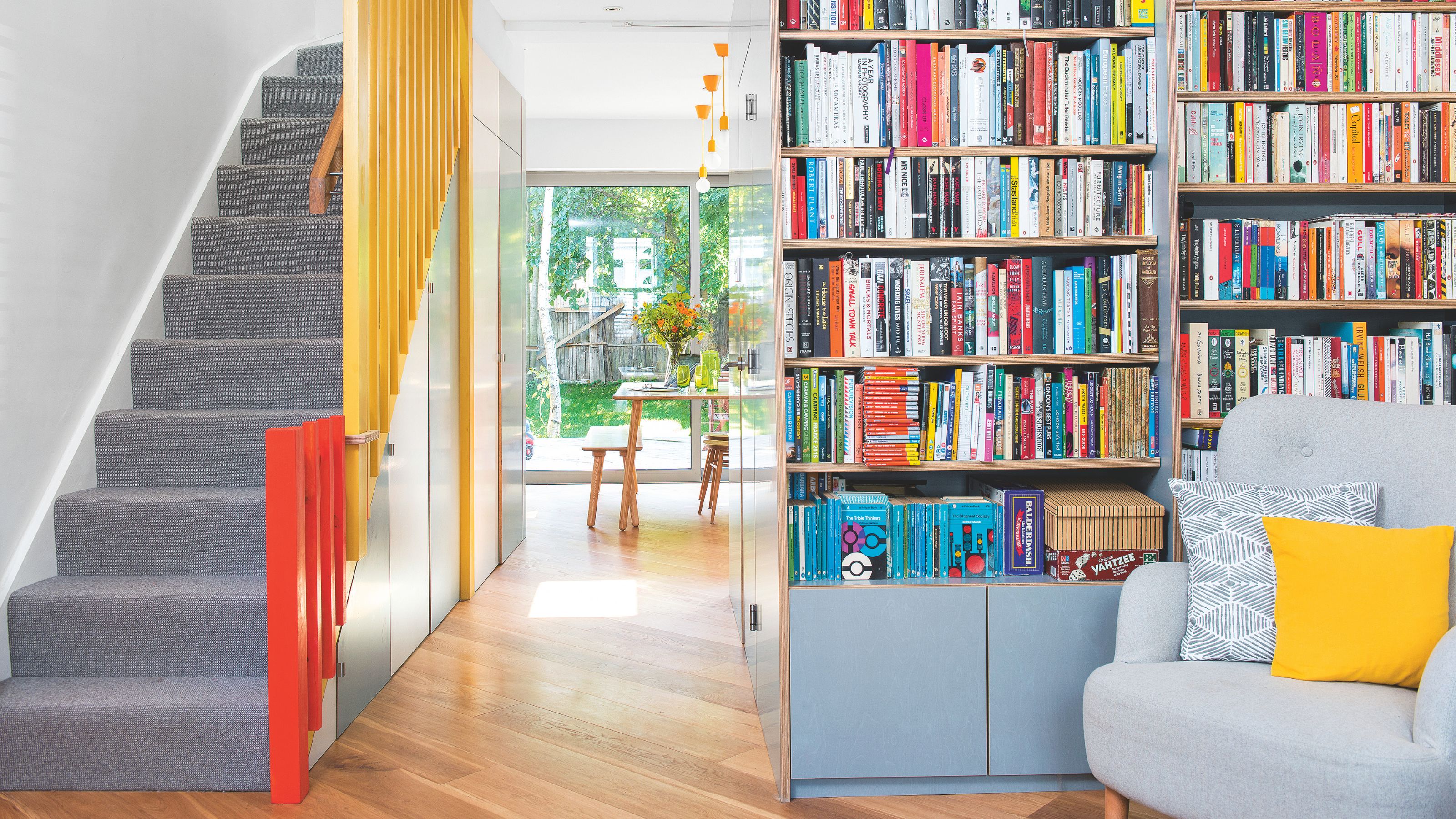 Grey carpeted staircase with red and yellow handrail, next to a large bookcase filled with books and a grey reading chair in front of it