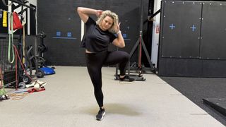 Woman demonstrates oblique crunch exercise in gym. She balances on one leg with her hands behind her head and elbows pointing to the side. She has raised her left knee out to the left side and bent her torso so her left elbow is close to her left knee.