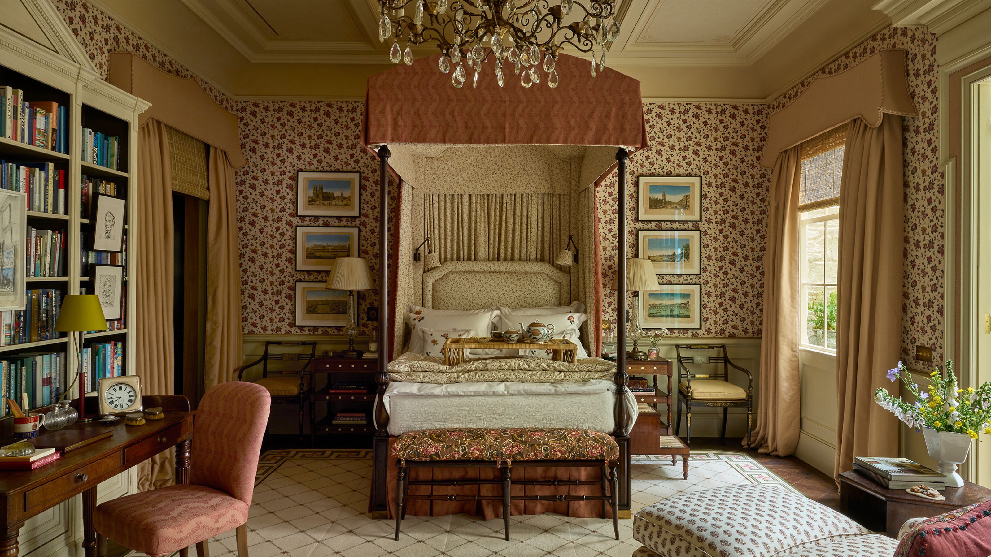 A traditional, elegant bedroom featuring a grand four-poster canopy bed, floral wallpaper, a crystal chandelier, and an ornate wooden desk with a pink chair adjacent to a tall bookcase.