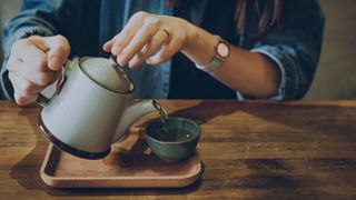 Woman pouring green tea into ceramic mug from wooden board