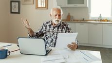 An older man looks annoyed and confused while looking at paperwork at his kitchen table.