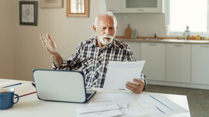 An older man looks annoyed and confused while looking at paperwork at his kitchen table.