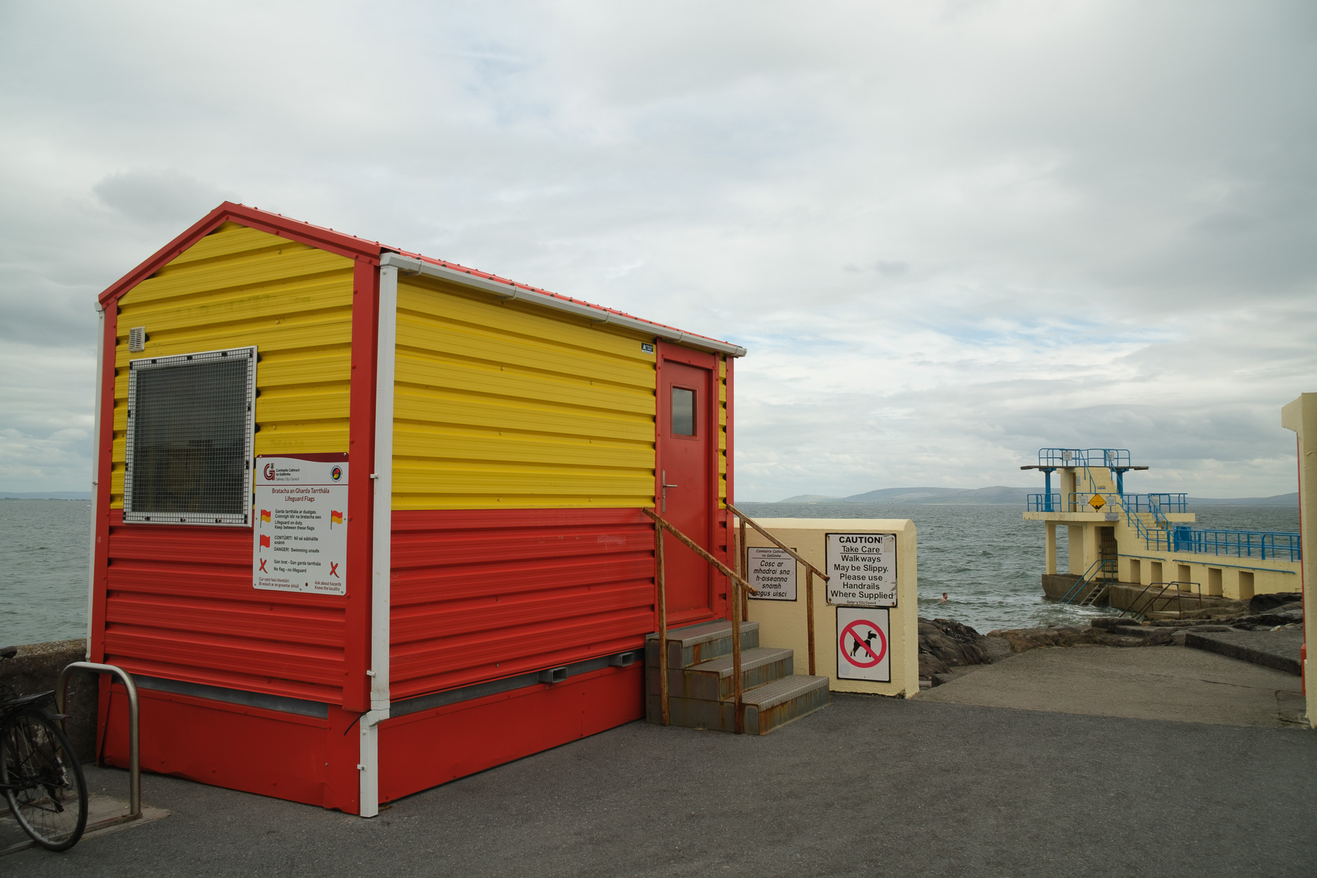 A brightly colored red and yellow lifeguard hut overlooks the sea, with a diving tower in the distance.