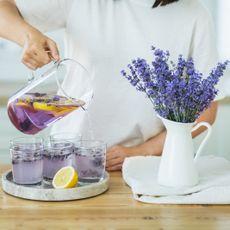 A woman pours lavender tea into five small cups next to a jar of honey and a pitcher of lavender flowers