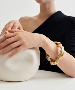 A headless image of a caucasian female model wearing a one should black ribbed dress with her hands graciously placed on a ceramic ball wearing a brass bangle on her left wrist