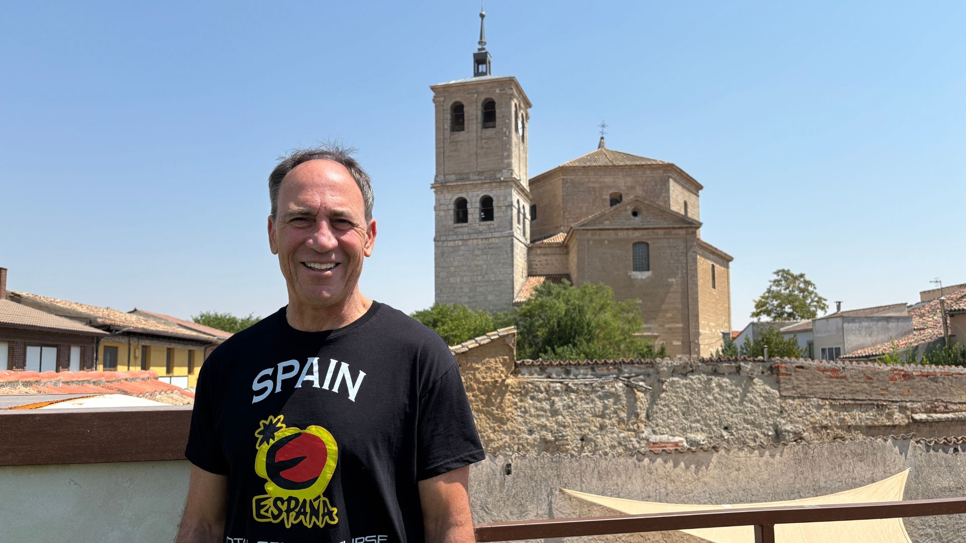 A man is photographed smiling as he stands in front of a church under blue skies on a summer day.